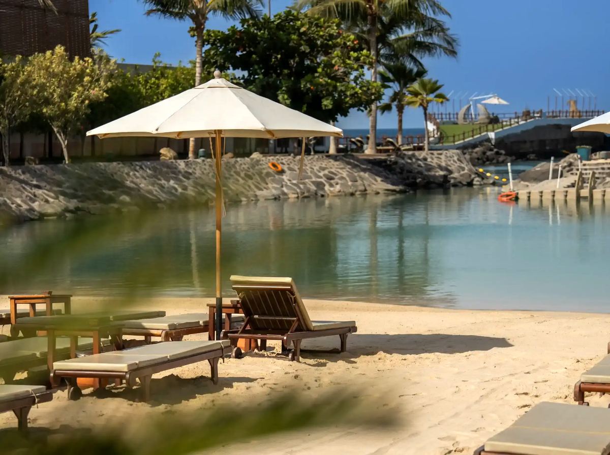 A white parasol and wooden sunloungers on soft sand beside a calm lagoon at La Maison D’Obhur