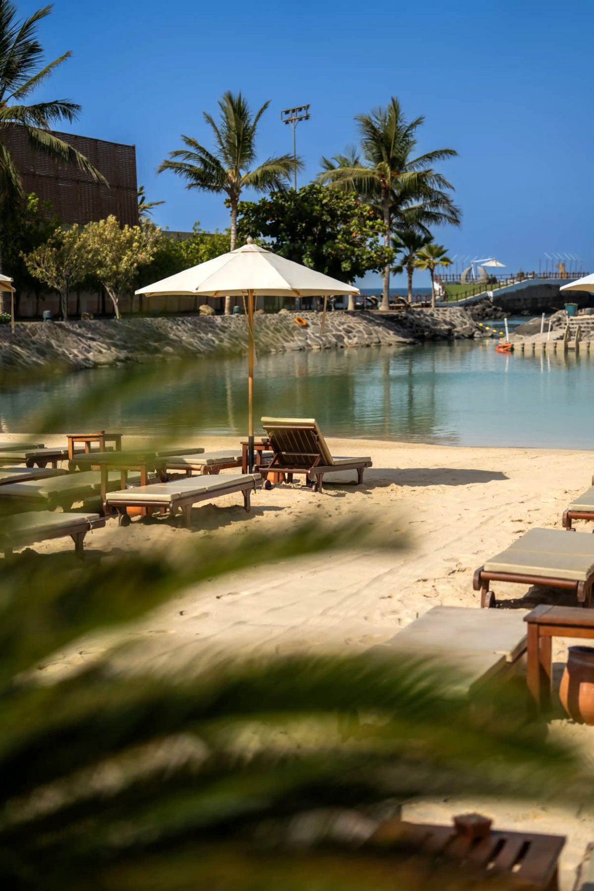 Lagoon beach with cushioned loungers and a parasol, framed by palm fronds, at La Maison D’Obhur