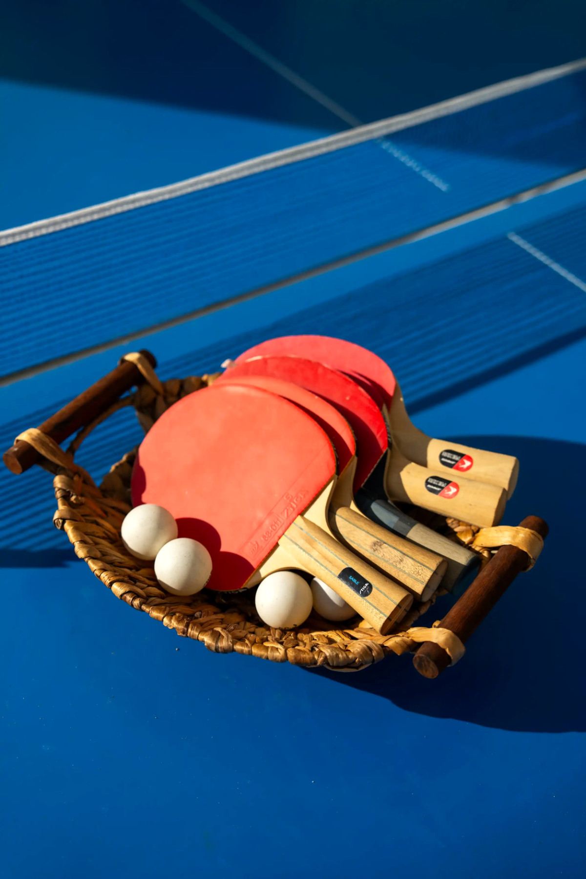 Table tennis paddles and balls in a woven basket on a blue table at Le Pont de l’Amitié