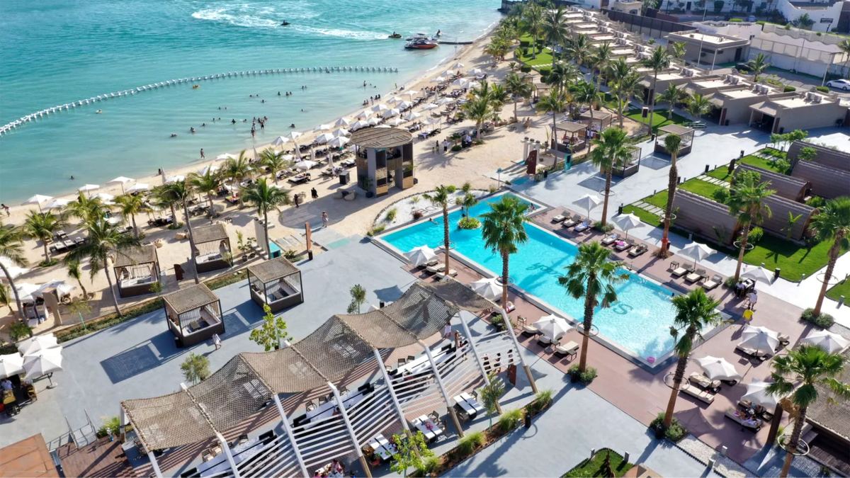 Aerial view of Shades Beach Club with its pool, shaded cabanas, palm trees, sandy beach and roped-off swimming area