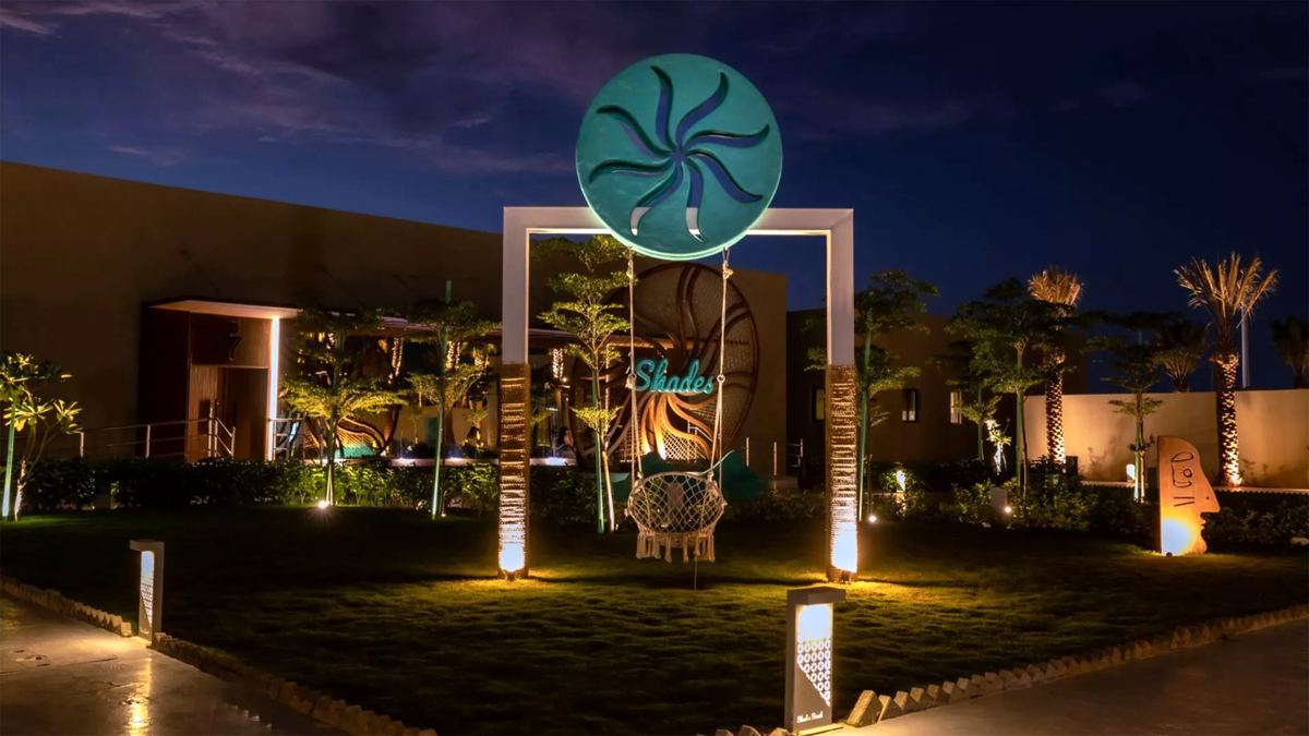 Shades Beach Club’s lit entrance arch with sunburst logo and a hanging swing at night
