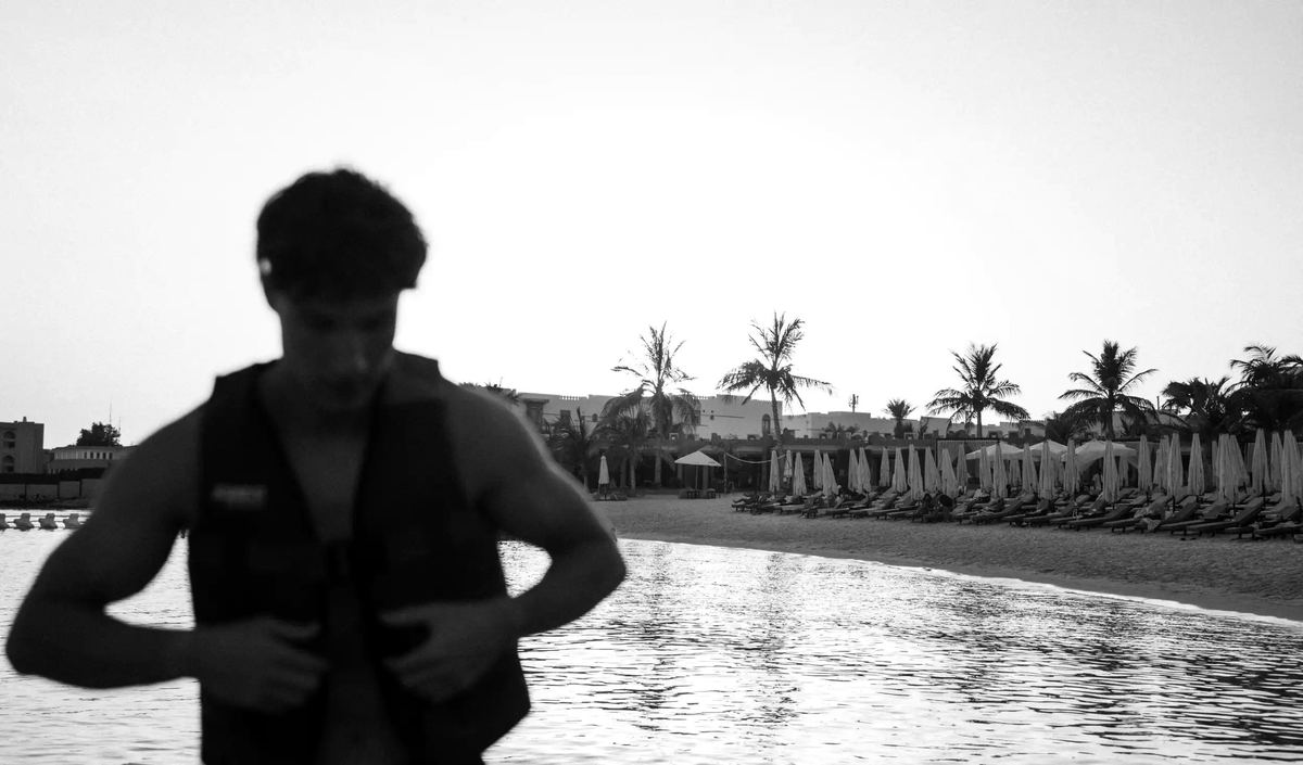 In black and white, a guest fastens a life vest by the shore at Shades Beach Club